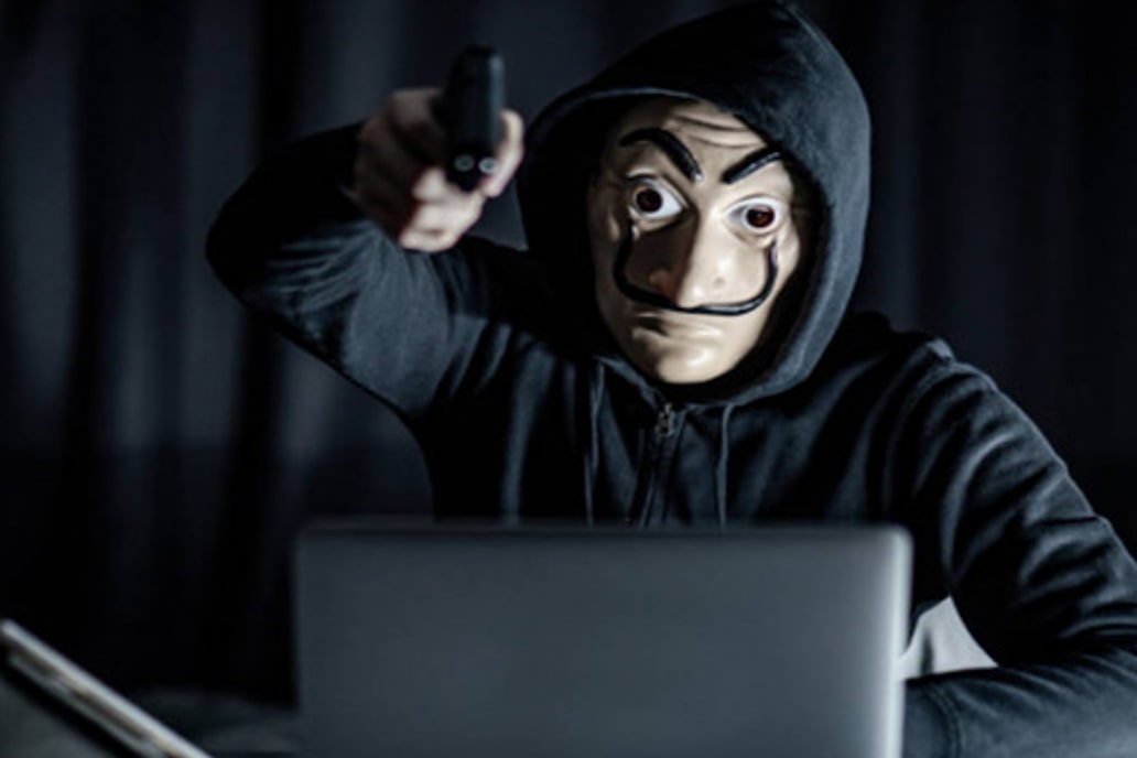 Un pirate informatique est assis, masqué, derrière un ordinateur, une arme à la main