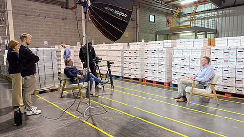 Sven wordt geïnterviewd in een magazijn met archiefdozen, terwijl de cameracrew het gesprek vastlegt.