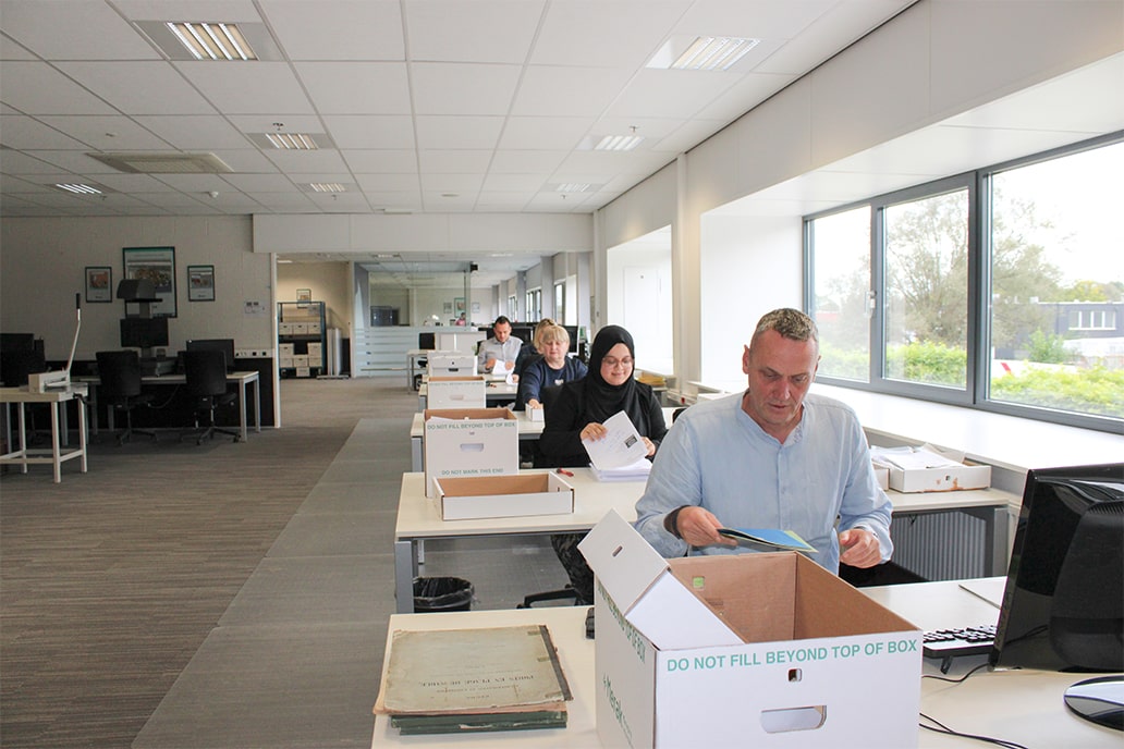 Een gefocust team sorteert documenten in dozen in een licht kantoor. Natuurlijk licht stroomt binnen via grote ramen. Efficiënt en ruim opgezet.